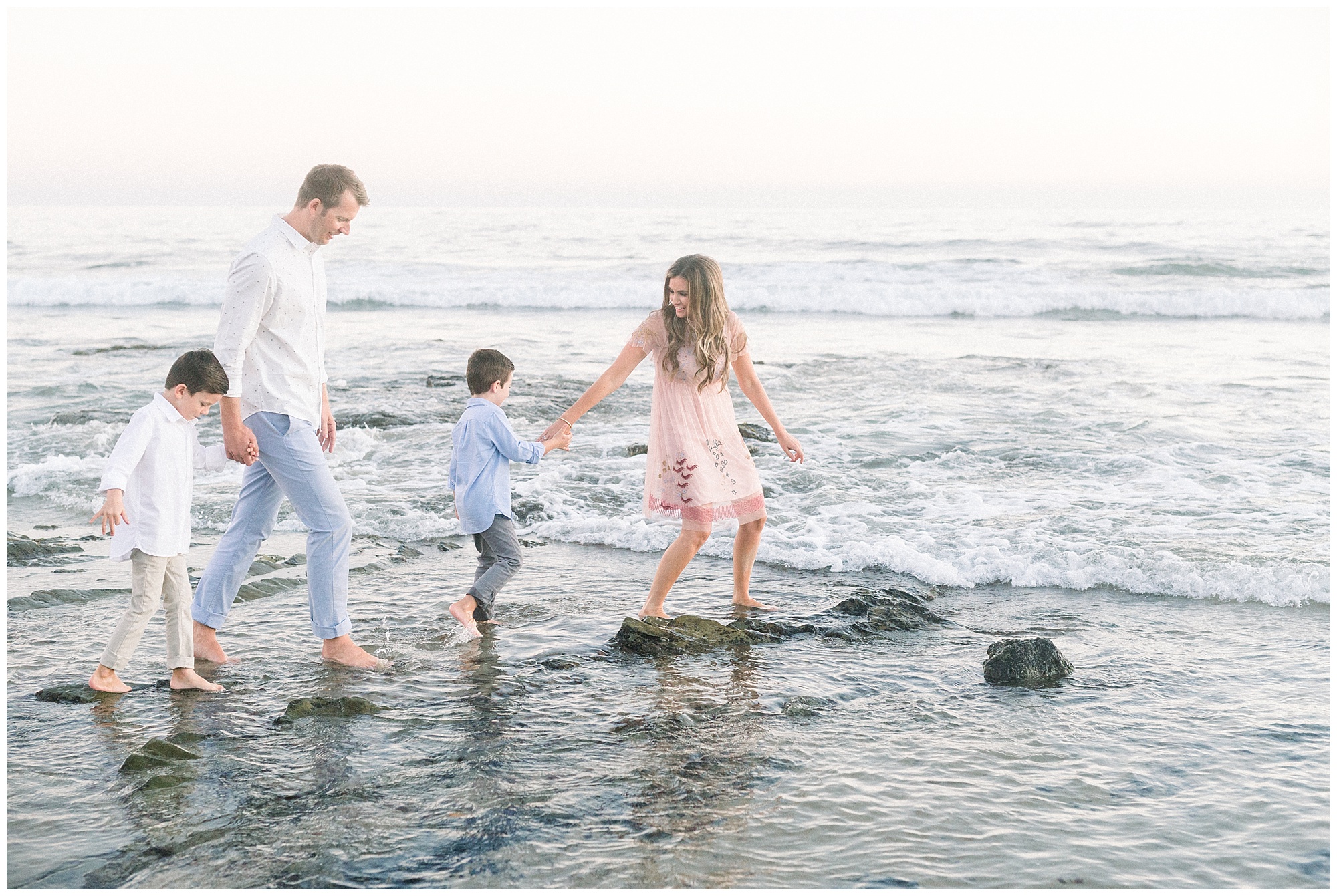 Family of four in neutral pink and blue colors photo in ocean water with rocks